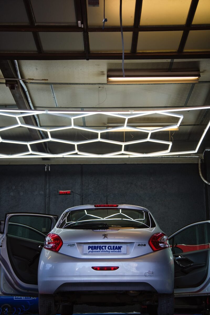 Vue intérieure de l'atelier : Peugeot 208 centrée sous la rampe hexagonale LED, portières arrière ouvertes en symétrie