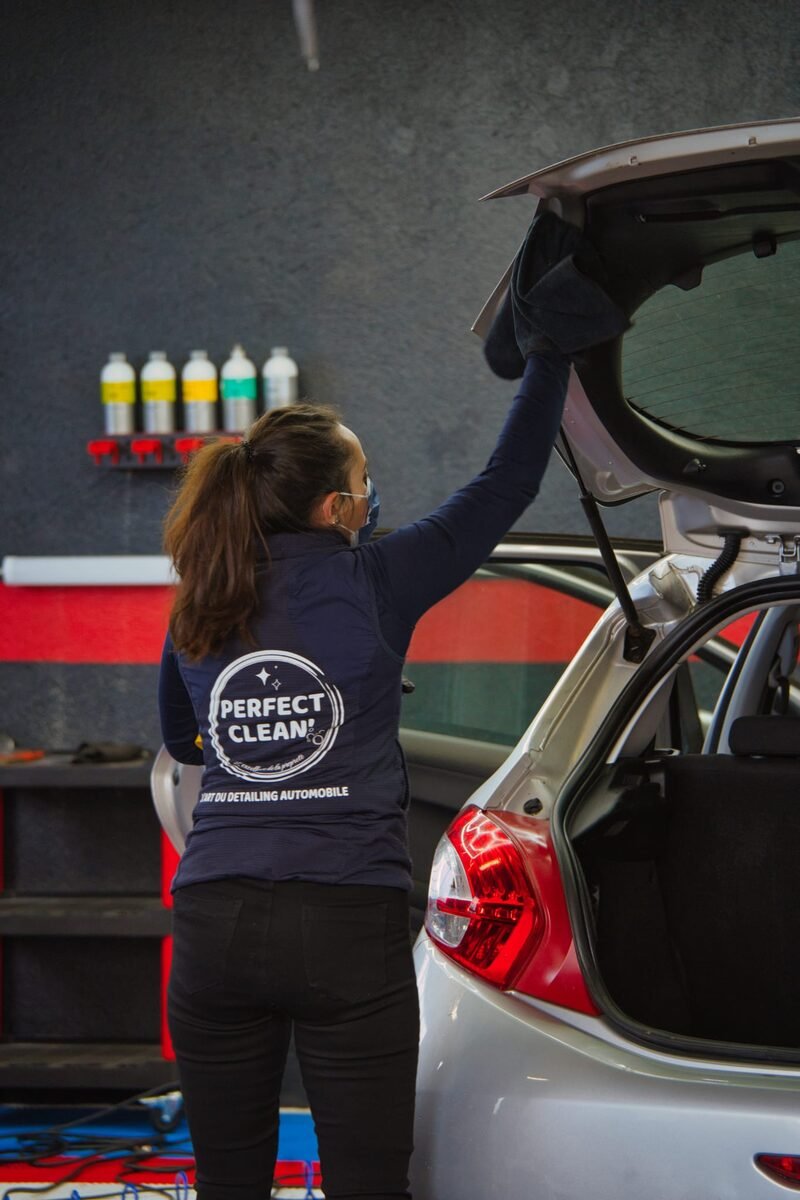 Détailleuse de dos essuyant le hayon, logo 'L'art du detailing automobile' visible sur la veste
